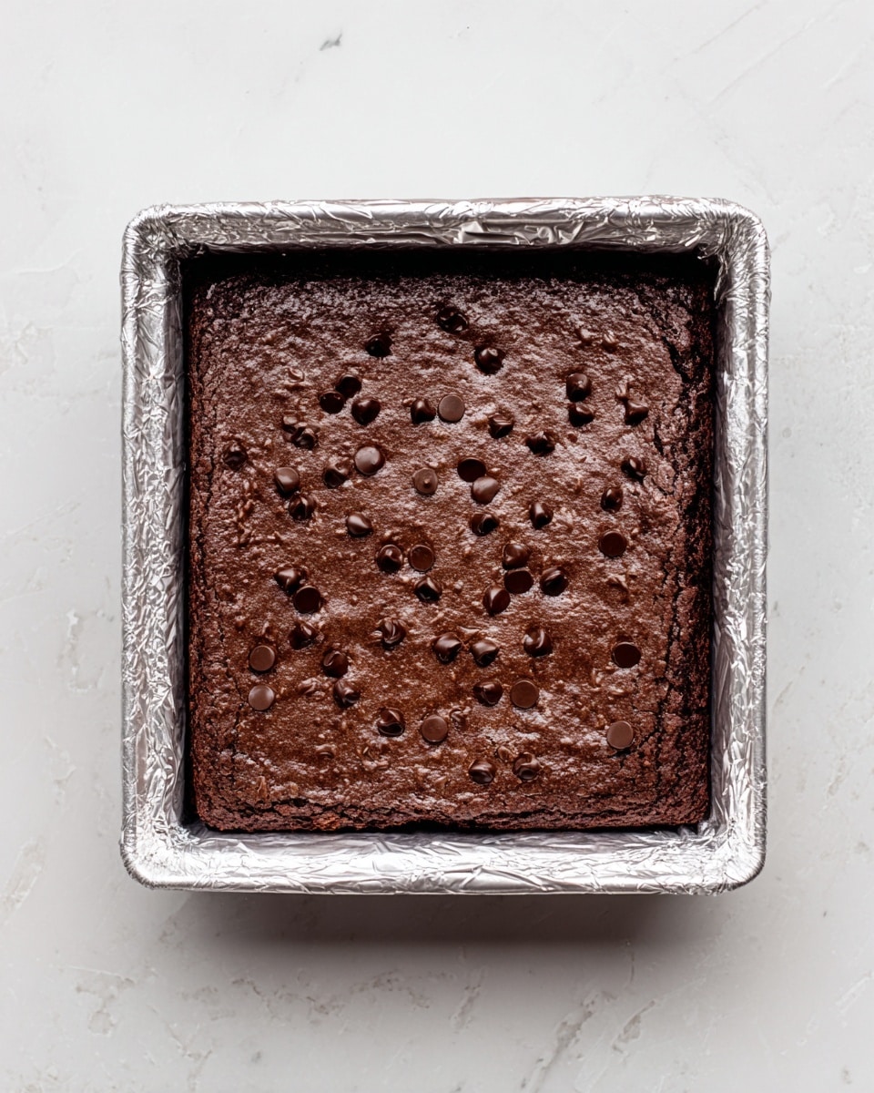 A square chocolate brownie with a dark brown color sits inside a foil-lined baking tray. The top layer is slightly textured with small, scattered shiny chocolate chips evenly spread across the surface. The tray rests on a white marbled texture background, creating a clean and bright setting. The brownie fills the tray completely, showing its soft and moist texture. photo taken with an iphone --ar 4:5 --v 7