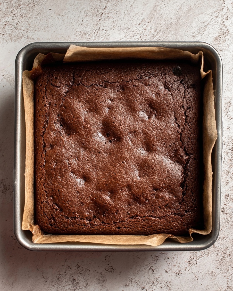 A square chocolate cake with a rich dark brown color sits in a metal baking pan lined with parchment paper, showing the cake’s slightly uneven, bumpy surface texture with small cracks and air pockets throughout. The pan rests on a white marbled textured surface, emphasizing the warm tone of the freshly baked cake. The edges of the cake have pulled slightly away from the paper, revealing its thickness inside the pan. Photo taken with an iphone --ar 4:5 --v 7