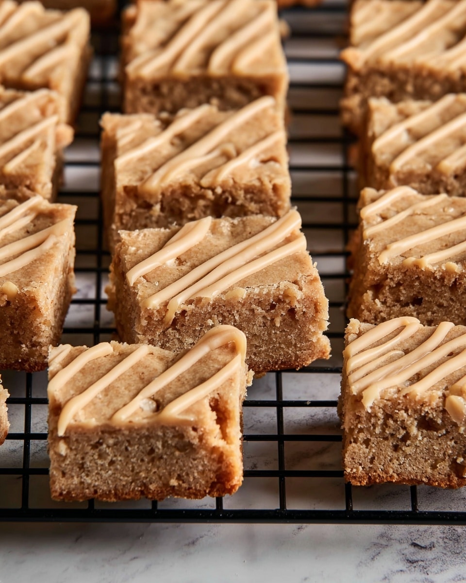 The image shows a close-up view of light brown square blondies placed in rows on a black wire cooling rack. Each blondie has one layer with a soft, moist texture filled with small bits that add a grainy look. Some of the blondies have a light tan drizzle in thin, wavy lines on top, adding a smooth contrast to the crumbly surface. The cooling rack sits on a surface with a white marbled texture, which softly contrasts with the warm tones of the blondies. The photo focuses tightly on the blondies, showing detailed texture and coloring. Photo taken with an iphone --ar 4:5 --v 7