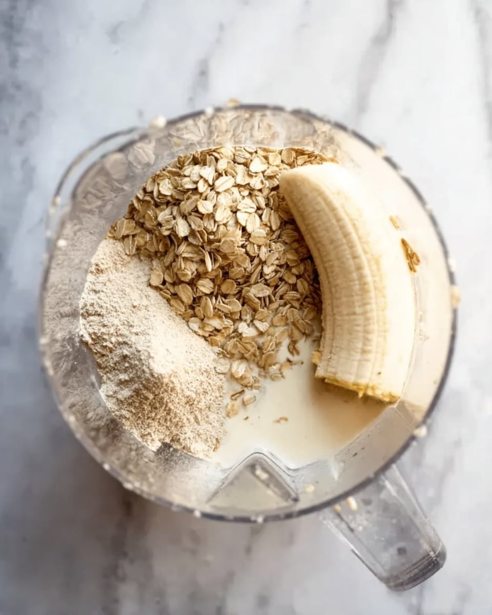 Inside a clear blender container placed on a white marbled surface, there are three main layers of ingredients. On the left side, there is a textured pile of light brown rolled oats, next to a smaller mound of beige powder. To the right, a whole peeled banana rests on top of a light cream liquid that pools at the bottom, with some soaked oats floating in it. The layers show a mix of dry and wet textures, ready to be blended. photo taken with an iphone --ar 4:5 --v 7