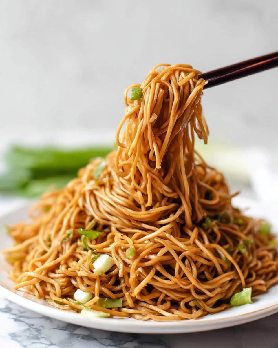 A large pile of thin brown noodles sits on a white plate with some small pieces of light green and white vegetables mixed in, mostly near the edges and under the noodles. A pair of dark brown chopsticks holds a clump of the noodles lifted above the plate, showing the smooth and slightly shiny texture of the noodles. The background is a white marbled surface with a blurry green vegetable in the back. Photo taken with an iphone --ar 4:5 --v 7