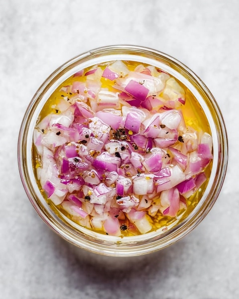 A clear round glass bowl holds a mixture of finely chopped red onions, mostly pinkish purple with some white parts, floating in light yellow oil with a shiny, smooth texture. The onions are evenly spread and partially submerged in the oil, which reflects light softly. Small bits of black pepper are scattered on top, adding dark specks. The bowl is placed on a white marbled textured surface, showing depth through the transparent bowl and the layered colors inside. photo taken with an iphone --ar 4:5 --v 7