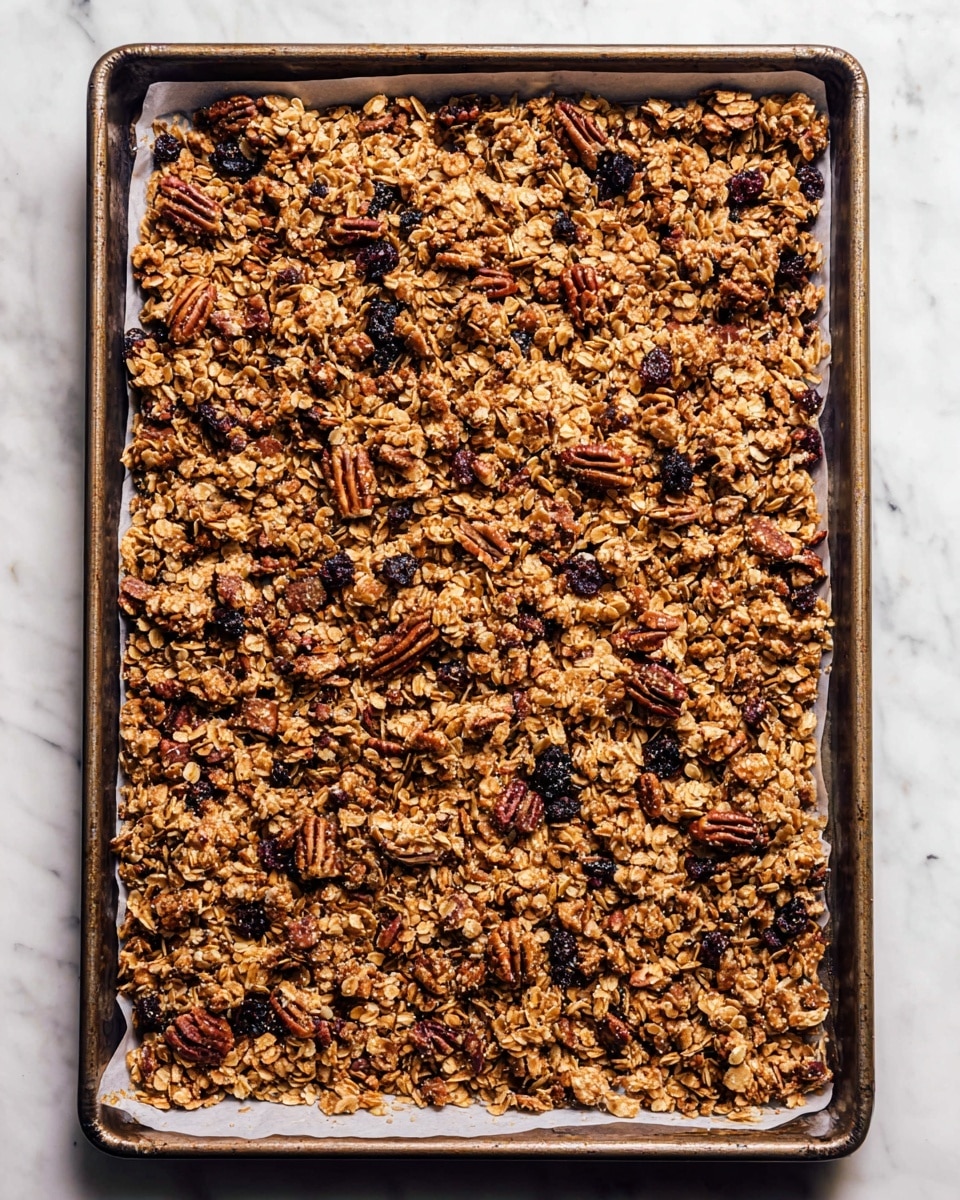 The image shows a large rectangular baking tray filled with a single thick layer of golden brown granola that includes oats, pecan nuts, and dark dried berries. The granola pieces vary in size and texture, some looking crunchy while others softer, evenly spread across white parchment paper lining the tray. The tray is resting on a white marbled surface, making the warm colors of the granola stand out clearly. photo taken with an iphone --ar 4:5 --v 7