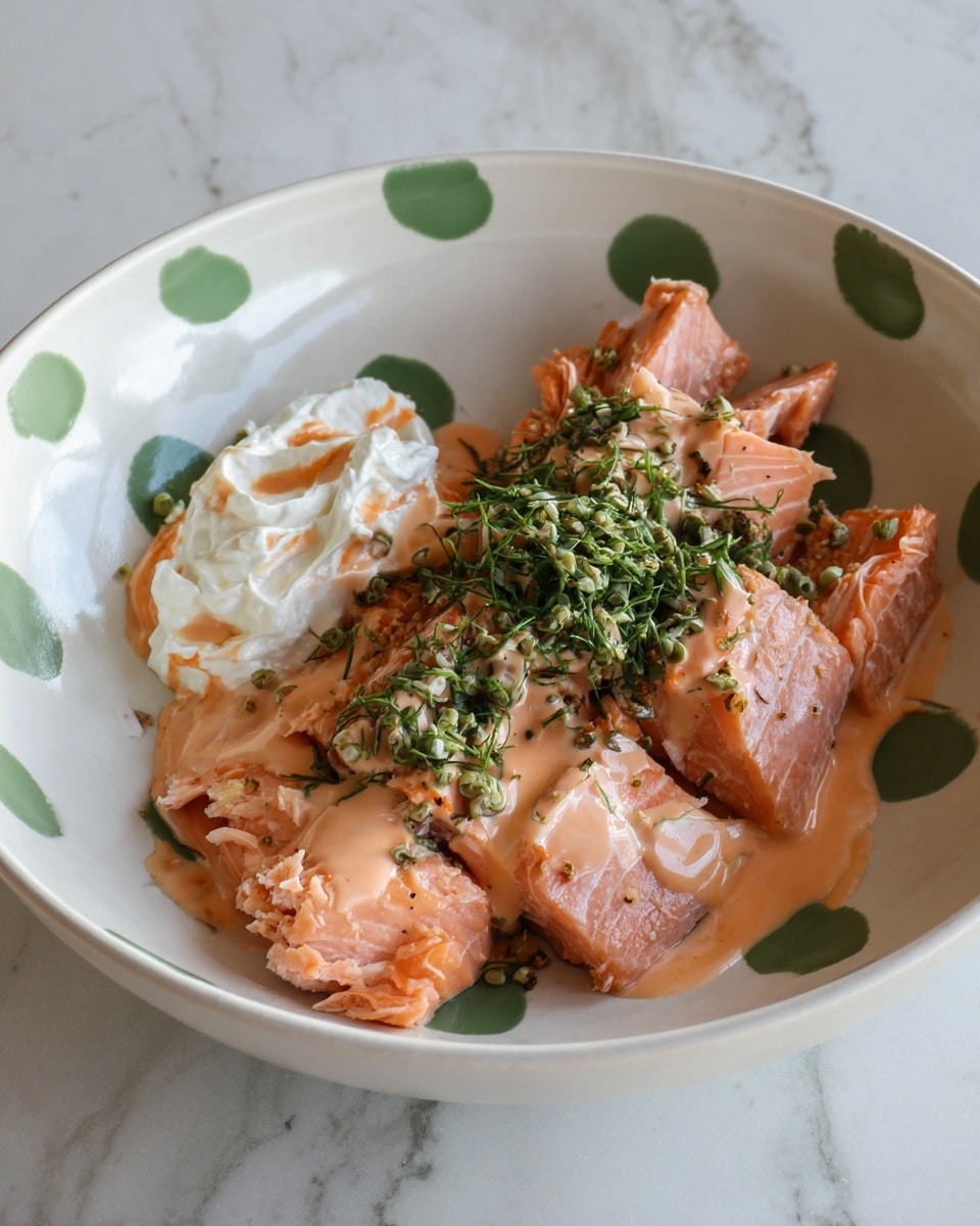 A white bowl with green dots holds several pieces of cooked pink salmon, layered roughly in the center. The salmon is topped with a pink-orange creamy sauce drizzled unevenly across the fish. On top of this sits a small pile of chopped green herbs, adding texture and color contrast. To the side, on the same white bowl, there is a dollop of whipped white cream or a similar soft white substance with a small drizzle of the same pink-orange sauce. The bowl is placed on a white marbled surface. photo taken with an iphone --ar 4:5 --v 7