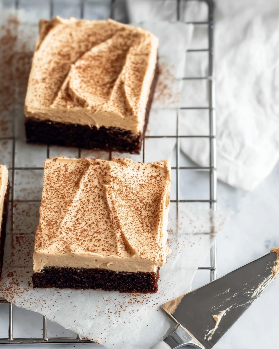 Two square pieces of dessert sit on a metal cooling rack above a white marbled surface. Each piece has two layers: a dark brown base layer and a thick, creamy beige frosting layer on top. The frosting is smooth with slight swirl textures and sprinkled evenly with fine brown powder. A metal spatula with some frosting on it rests nearby on the white marbled surface. The photo has soft natural lighting, showing details of the frosting’s texture and the cooling rack’s grid pattern photo taken with an iphone --ar 4:5 --v 7