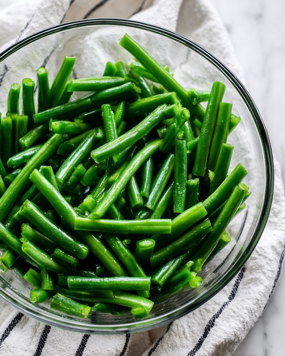 A clear glass bowl filled with fresh green beans that are cut into smaller pieces. The green beans look shiny and bright green, showing a smooth and firm texture. The bowl sits on a white marbled surface with a white cloth that has thin black stripes underneath it. photo taken with an iphone --ar 4:5 --v 7