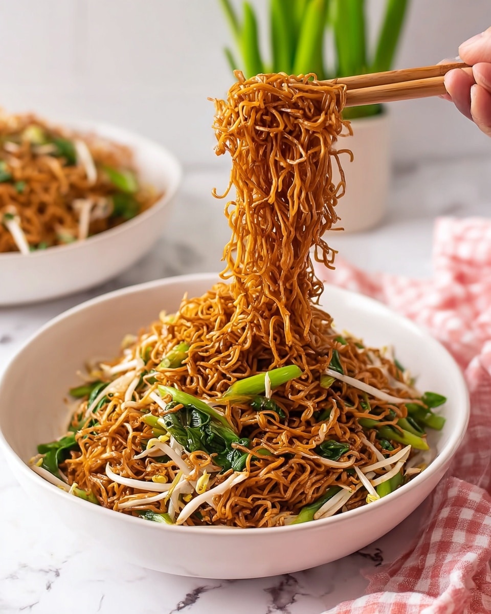 A large white bowl filled with a layer of thin, brown fried noodles mixed with light-colored bean sprouts and bright green leafy vegetables. A woman's hand holding wooden chopsticks lifts a portion of the noodles above the bowl, showing their curly and tangled texture. In the background, there is another white bowl containing more noodles, placed on a white marbled surface, with a small green plant nearby. A pink and white checkered cloth is partially visible at the bottom of the image. Photo taken with an iphone --ar 4:5 --v 7