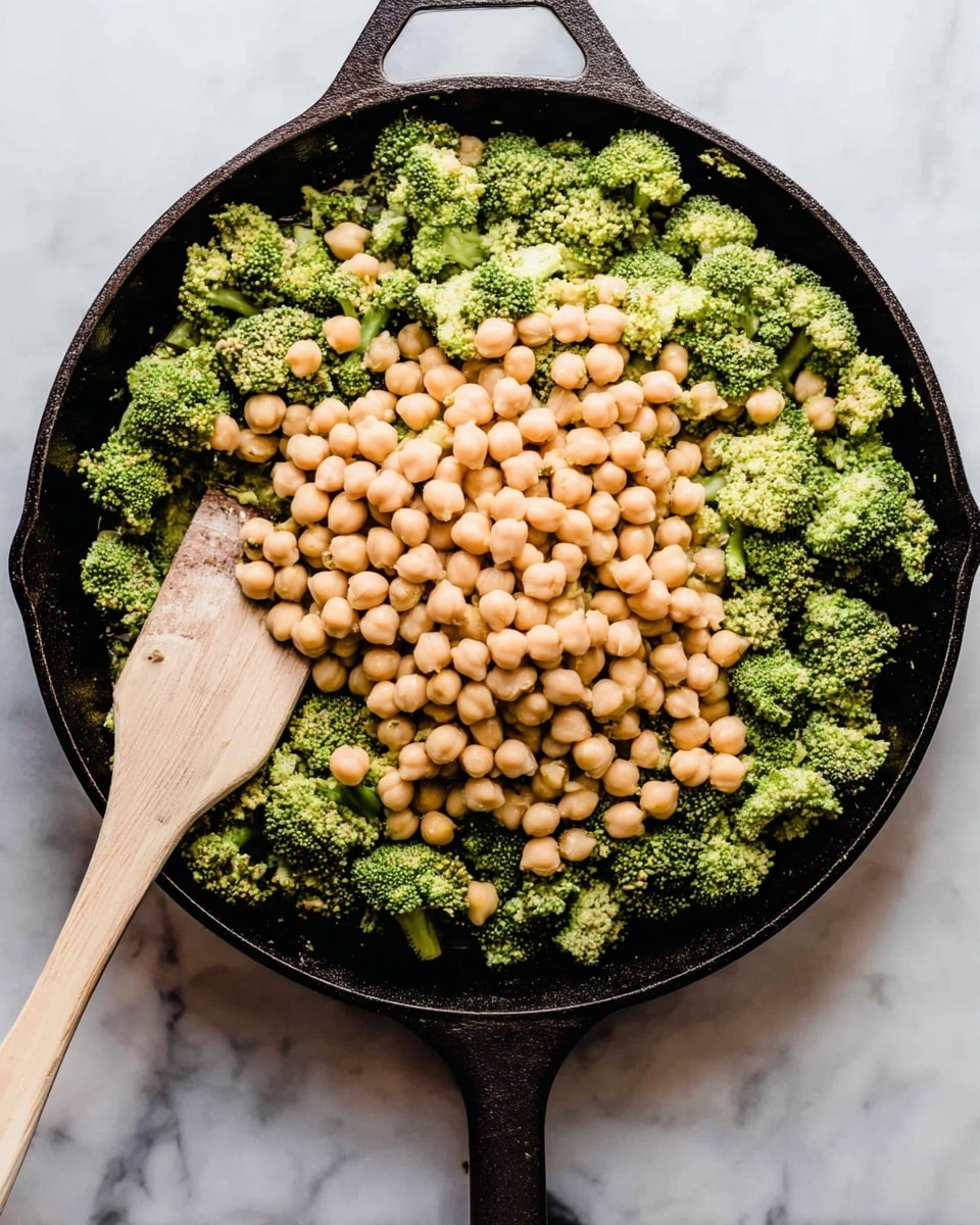 A black cast iron pan holding two main layers: a base layer of green broccoli florets with a textured, slightly rough surface, topped with a large pile of smooth, light beige chickpeas clustered in the middle. A wooden spatula with a light brown handle rests on the left side, partly touching the broccoli and chickpeas. The pan is set on a white marbled surface, visible around its edges. Photo taken with an iphone --ar 4:5 --v 7