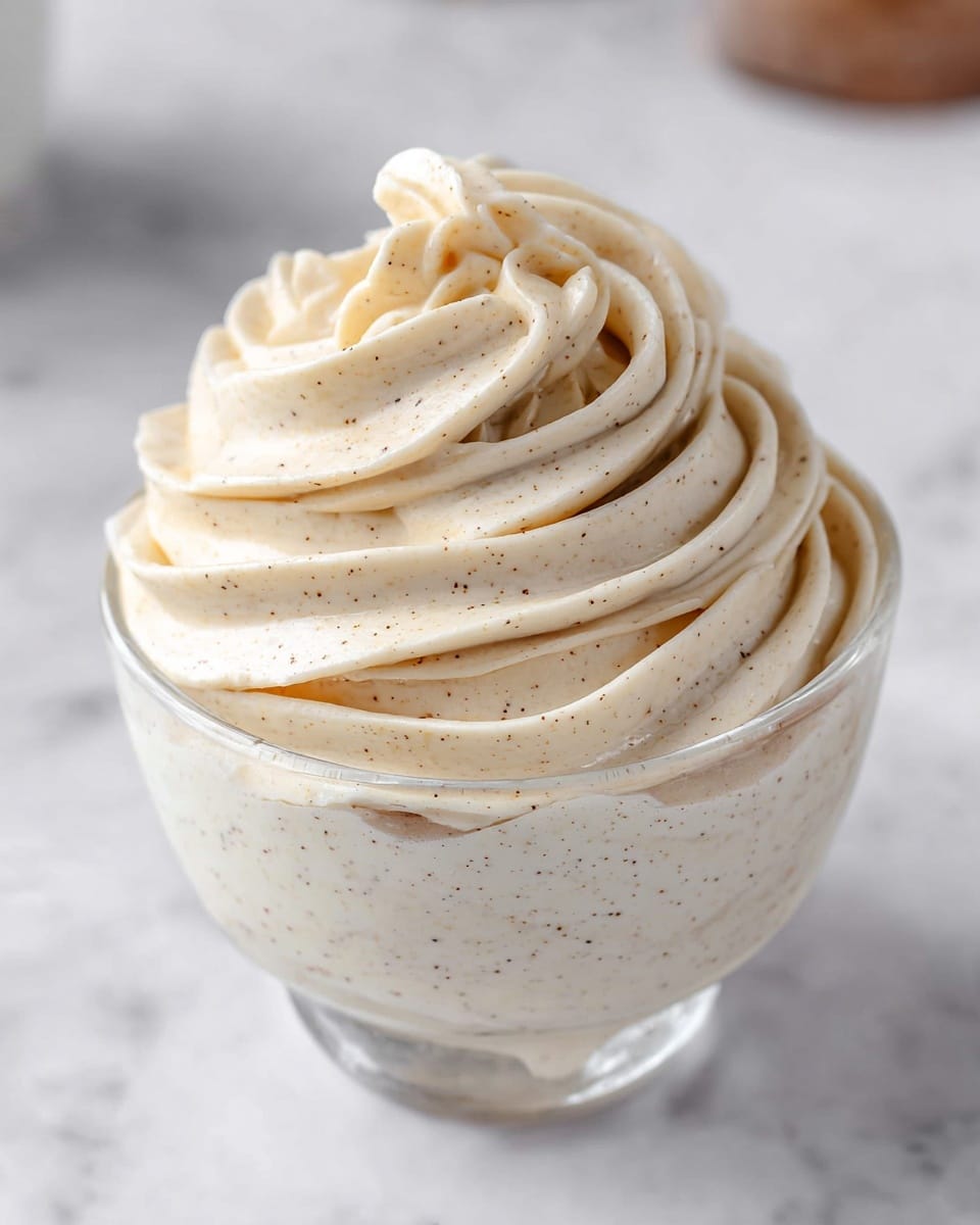 A clear glass dessert bowl filled with a thick swirl of light beige cream, dotted with tiny vanilla bean specks, piled high in soft, smooth spirals that create a slightly pointed peak at the top. The creamy texture looks rich and fluffy, with well-defined ridges from the piping. The bowl sits on a white marbled surface, with a blurred background that includes soft neutral shapes suggesting kitchen items. Photo taken with an iphone --ar 4:5 --v 7