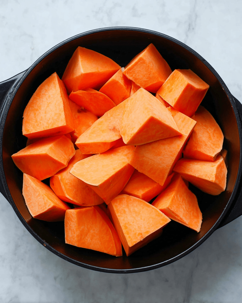 Chunks of bright orange sweet potatoes fill a black pan, each piece sliced into thick, uneven shapes with smooth, matte surfaces and sharp edges. The sweet potatoes are piled closely together, covering the pan completely, sitting on a white marbled surface. Photo taken with an iphone --ar 4:5 --v 7