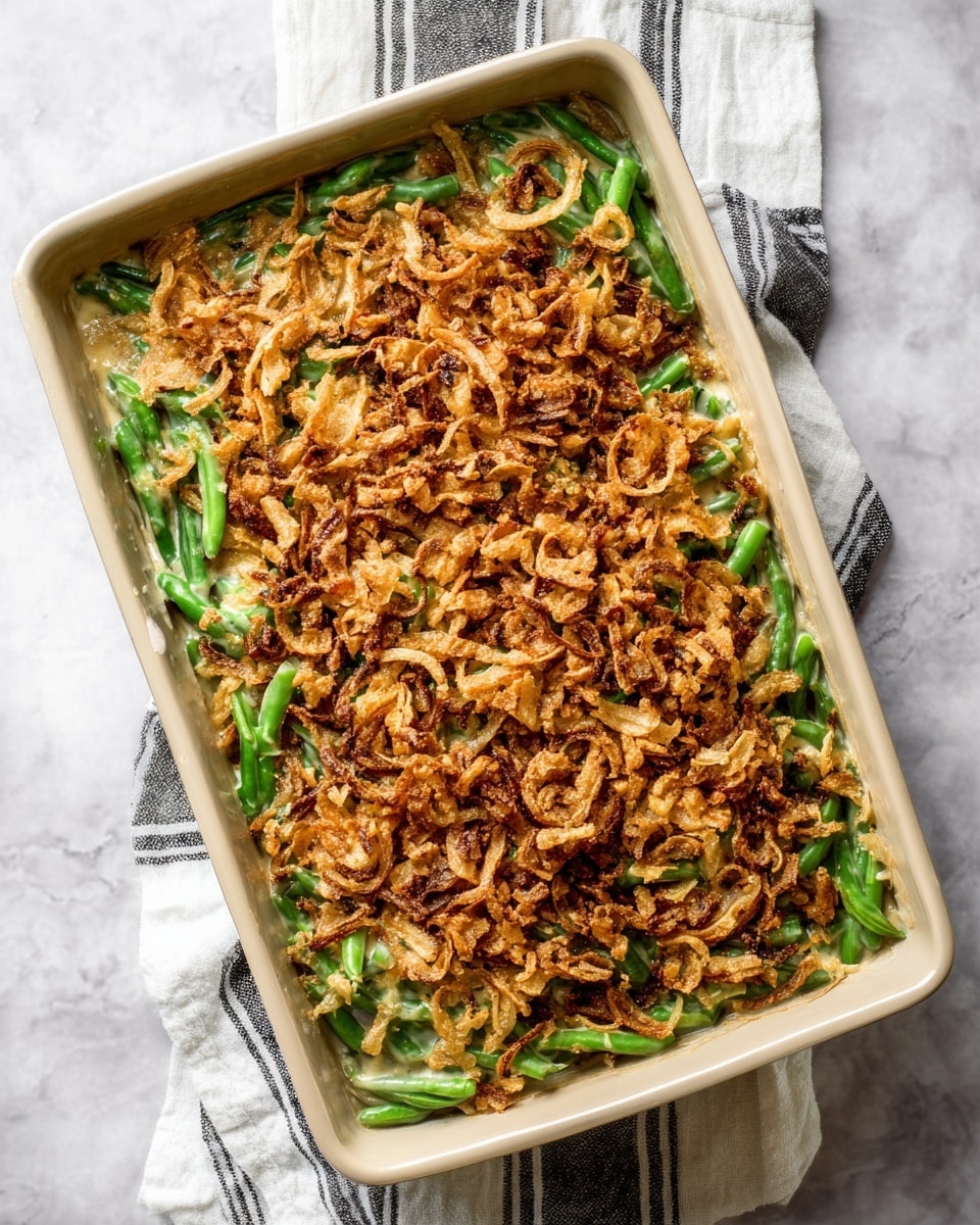A rectangular baking dish filled with a green bean casserole is shown from above. The dish has three visible layers: the bottom layer is bright green beans, the middle layer is a creamy light beige sauce that slightly peeks through the sides, and the top layer is a thick, crispy, golden-brown fried onion topping that covers the whole surface unevenly with some darker spots. The dish sits on a white cloth with thin black stripes on a white marbled textured surface. Photo taken with an iphone --ar 4:5 --v 7