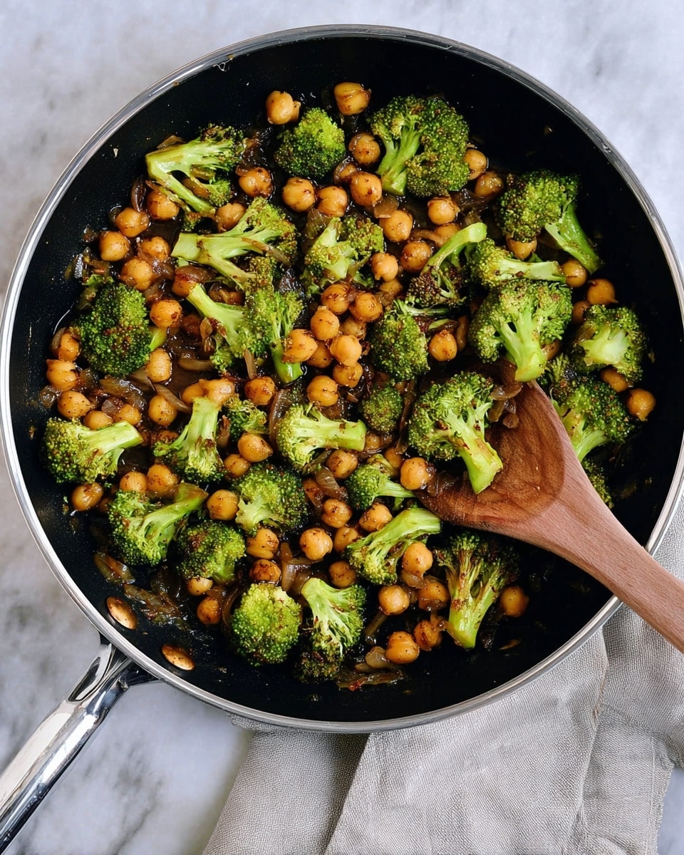 A black frying pan filled with cooked green broccoli pieces and round golden-brown chickpeas, both coated in a light brown sauce with small bits of cooked onion mixed in. A wooden spoon rests inside the pan, partially under some broccoli. The pan is placed on a white marbled surface with a light gray cloth nearby. Photo taken with an iphone --ar 4:5 --v 7