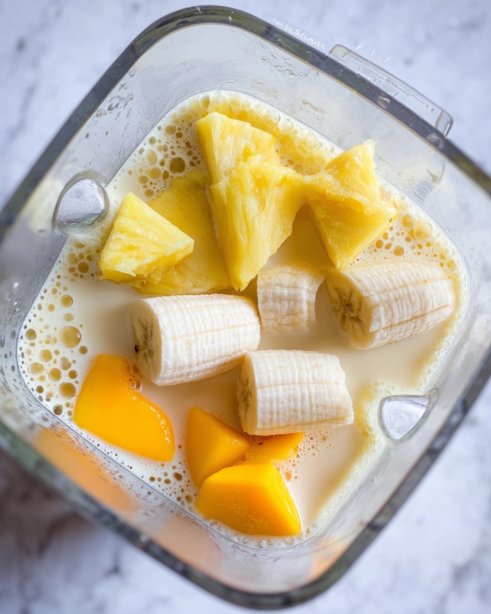 The image shows a blender container filled with pieces of white peeled banana, bright yellow pineapple chunks, and orange mango pieces immersed in a light creamy liquid base, likely a smoothie or juice mixture. The banana pieces are large and soft in texture, placed mainly on the right and bottom left side, while the pineapple chunks sit near the top left corner with a fibrous texture. The mango chunks, showing a shiny and smooth surface, occupy the bottom center and right side. The white marbled texture surface is visible around the blender's edges. photo taken with an iphone --ar 4:5 --v 7