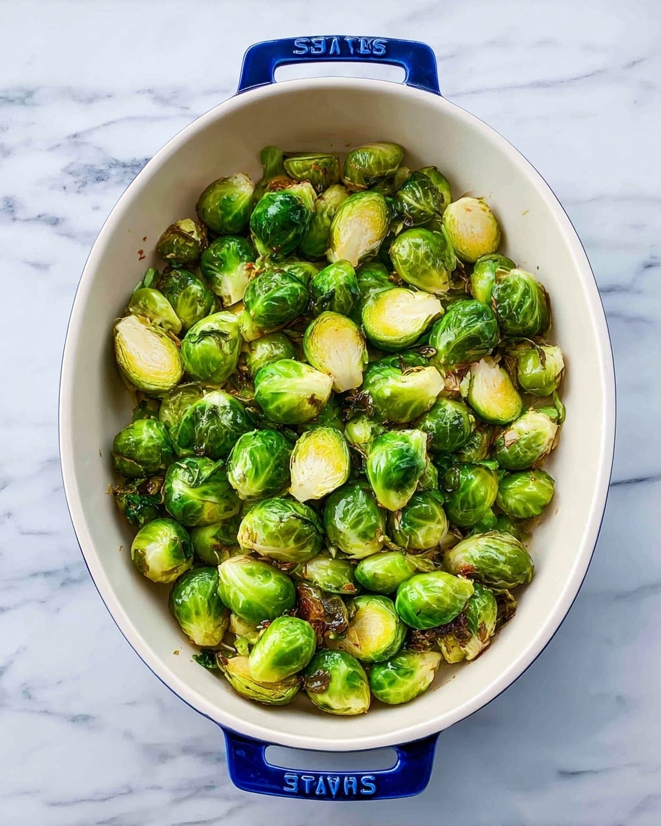 The image shows a white oval baking dish with blue handles on both sides, filled with roasted Brussels sprouts. The Brussels sprouts are halved and whole, bright green with some slightly browned edges, giving a mix of soft and crispy textures throughout. The dish is placed on a white marbled surface, and the Brussels sprouts are spread evenly, covering the entire base of the dish. photo taken with an iphone --ar 4:5 --v 7