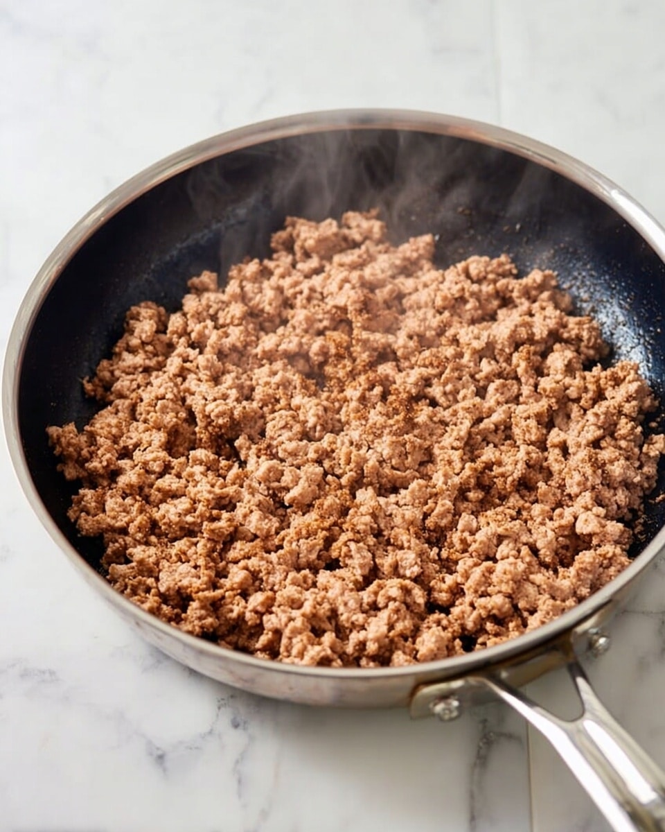 A close-up view of cooked ground meat in a shiny silver pan with a black interior, spread evenly across the pan's surface showing a crumbly texture with a light brown color. The pan sits on a white marbled surface, and steam rises gently from the meat, indicating it is hot. The pan handle extends to the right with a reflective silver finish. Photo taken with an iphone --ar 4:5 --v 7