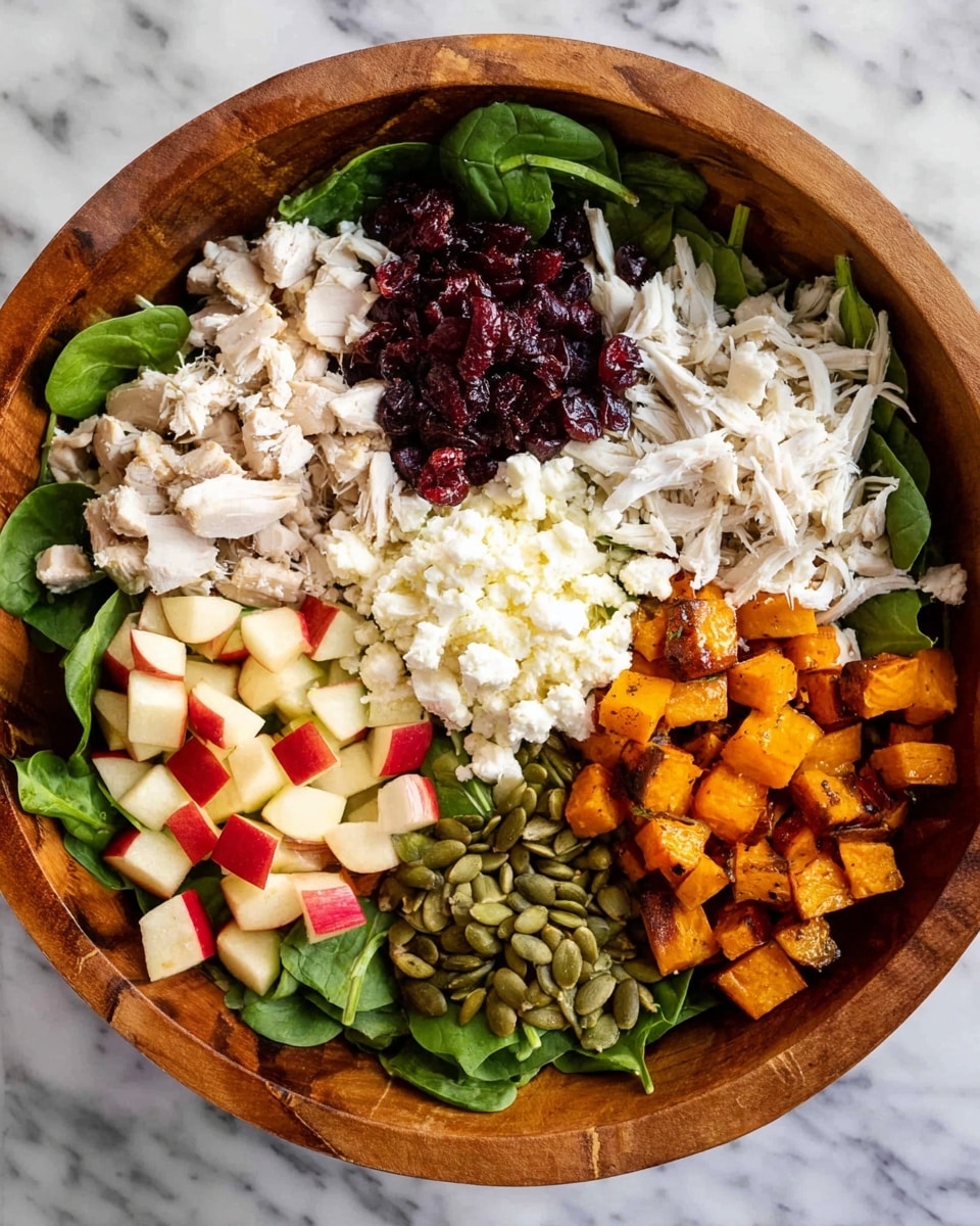 A wooden bowl holds a colorful layered salad arranged in sections. The base is fresh green spinach leaves spread evenly along the bottom. On top, there are diced red apples with smooth, shiny skin on one side. Next to the apples, there are small dark red dried cranberries that look slightly wrinkled. Adjacent to that is a pile of white shredded chicken with soft, stringy texture. In the center, a mound of white crumbly cheese adds a creamy contrast. On the opposite side, cubed roasted sweet potatoes with orange and brown edges add a rich, roasted look. Lastly, green pumpkin seeds sit near the bottom, adding a smooth, matte finish. The bowl is placed on a white marbled surface. photo taken with an iphone --ar 4:5 --v 7