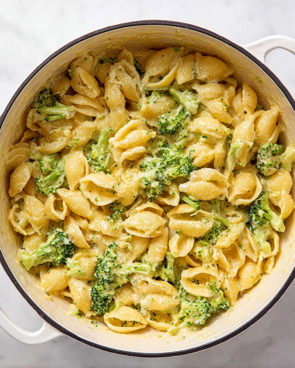 The image shows a white pot filled with a creamy pasta dish. The pasta is shell-shaped and covered in a smooth, light yellow cheese sauce mixed evenly throughout. Bright green broccoli pieces are scattered across the pasta, adding a fresh contrast of color. The pot sits on a white marbled surface, enhancing the colors of the food. The texture of the pasta looks soft and the sauce coats every piece well with small bits of broccoli visible in and around the shells. Photo taken with an iphone --ar 4:5 --v 7