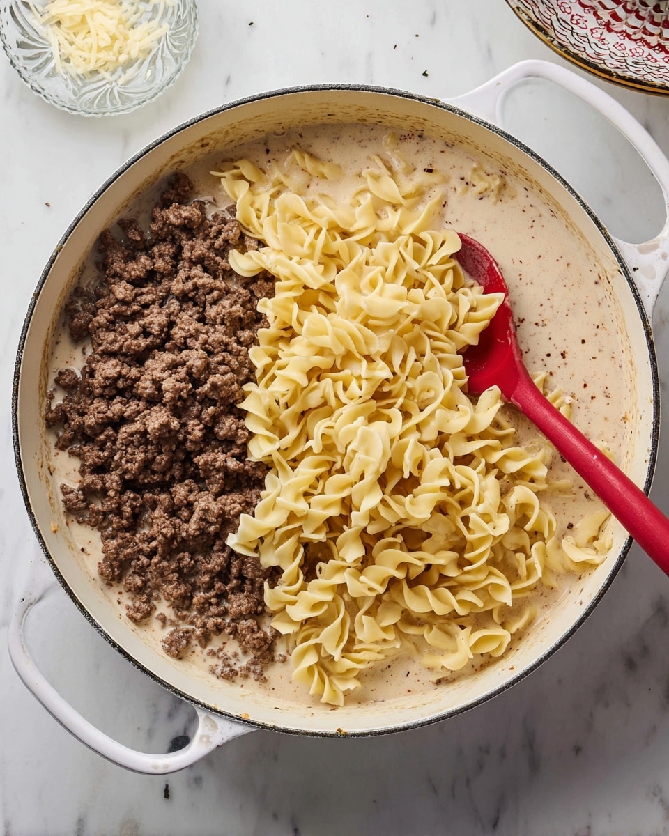 In a white pan with two handles, the bottom layer shows a creamy, light beige sauce with small specks of seasoning. On top of this sauce, there are two distinct layers side by side: on the left, cooked ground beef in small brown crumbles, and on the right, pale yellow cooked curly egg noodles, slightly shiny from the sauce. A red spoon is partially submerged in the noodles and sauce, resting on the right side of the pan. The pan is placed on a white marbled surface with some small dishes partially visible around it. Photo taken with an iphone --ar 4:5 --v 7