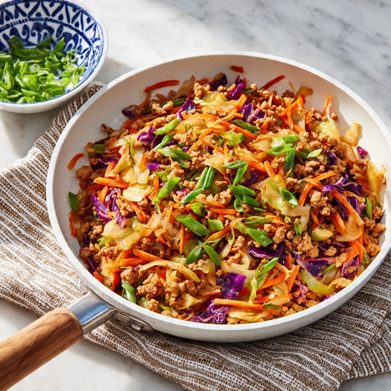 A white plate with a thin black rim holds a colorful stir-fry dish made of finely ground brown meat mixed with thin strips of bright orange carrots, pale yellow cabbage, and small pieces of purple cabbage. The dish is topped with small slices of light green scallions. A black-handled fork with a gold head rests on the right side of the plate. The plate sits on a white marbled surface, next to a beige and white striped cloth, with a small white bowl filled with chopped green onions visible in the upper left corner. Photo taken with an iphone --ar 4:5 --v 7