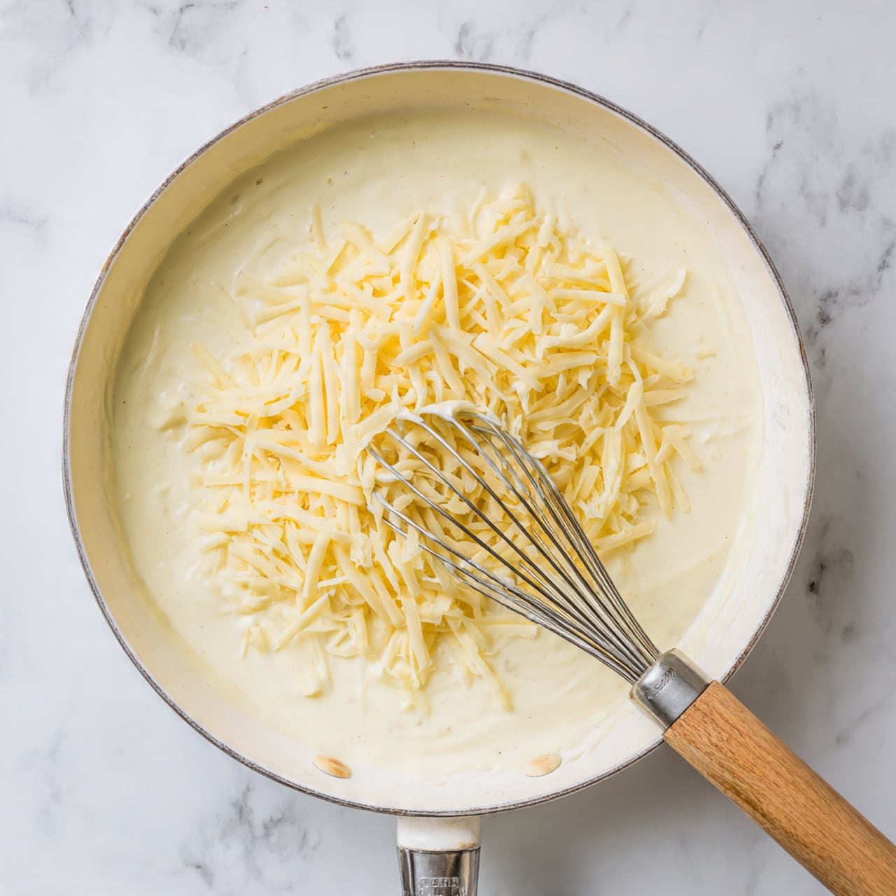 In a white cast iron skillet, creamy white sauce covers the base with a smooth texture, slightly thick. On top of the sauce, there is a pile of shredded light yellow cheese near the center-right. A metal whisk with a wooden handle is resting diagonally inside the skillet, partly touching the cheese and sauce. The skillet handle extends downward, and it sits on a white marbled surface. Photo taken with an iphone --ar 4:5 --v 7