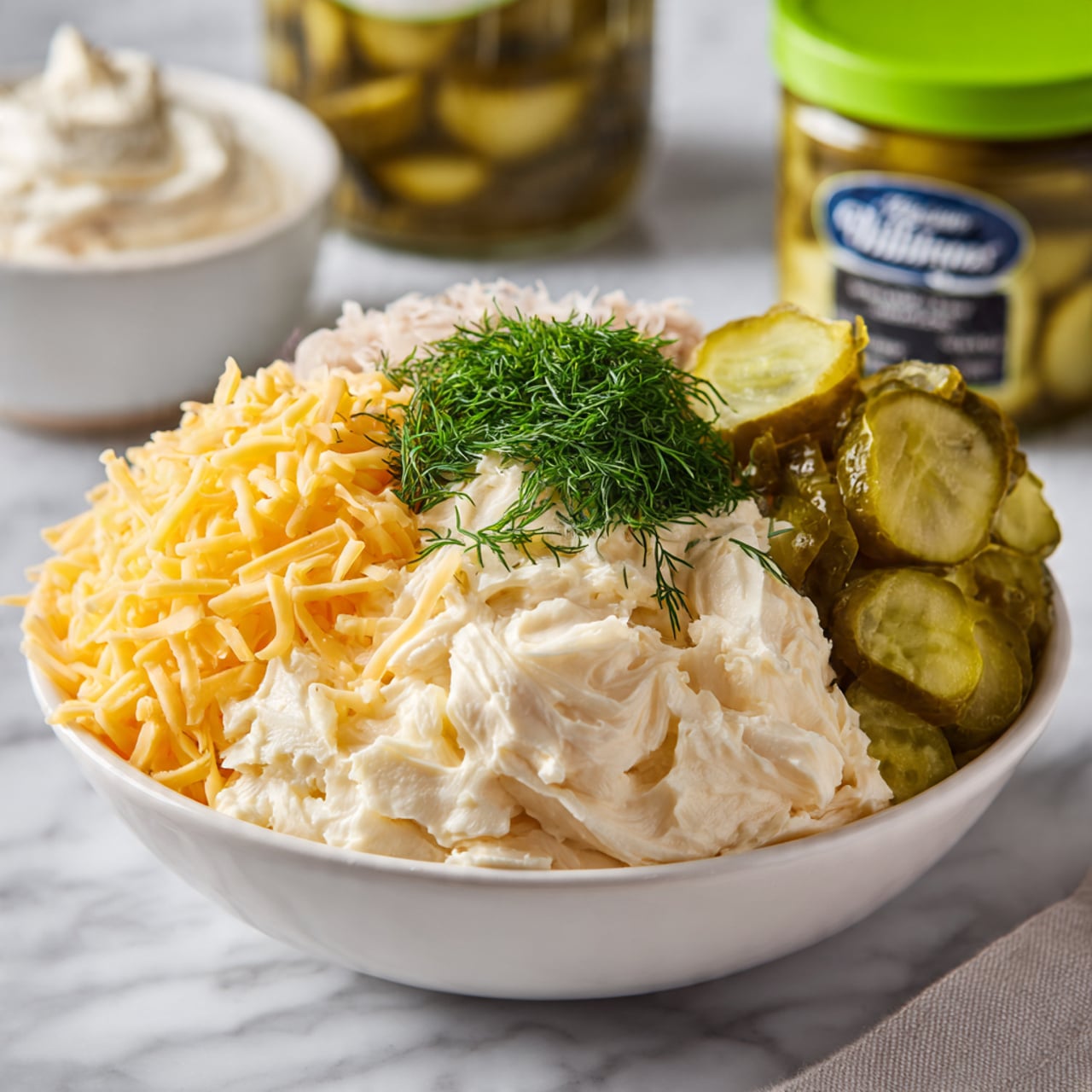 A white bowl sits on a white marbled surface filled with four distinct layers of ingredients ready to be mixed: at the bottom is a creamy, pale base; on the top left is a bright pile of shredded yellow cheddar cheese; to the right of that is a layer of finely chopped green pickles; and in the center front is a small mound of fresh, finely chopped dark green dill. In the background, there is a jar of pickles with a bright green lid and a container of sour cream, both slightly blurred. Photo taken with an iphone --ar 4:5 --v 7
