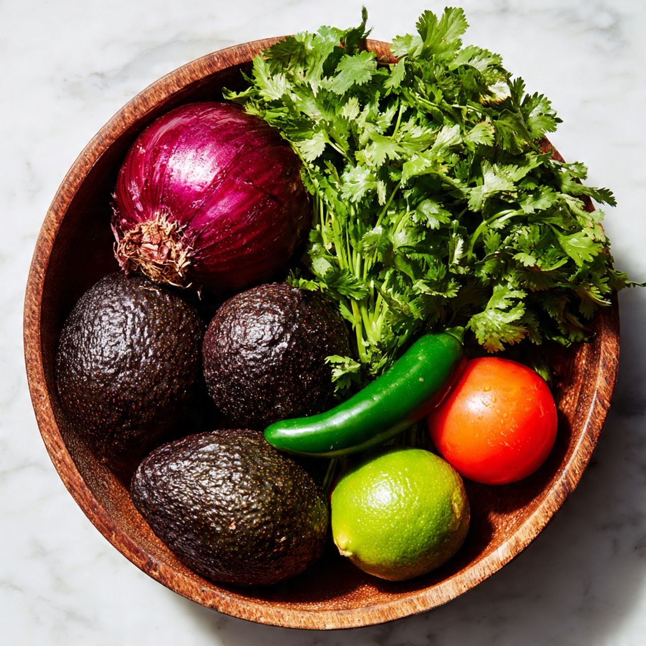 A round wooden bowl sits on a white marbled surface, filled with fresh ingredients arranged closely together. Inside the bowl, there are three dark green avocados with bumpy textures positioned around the bottom and left side. Above them is a large red onion with a smooth but cracked outer skin. To the right, a bunch of bright green leafy cilantro fills the space, with its stems visible. Two smooth, red tomatoes rest at the bottom right, next to a single shiny green lime and a dark green jalapeño pepper placed near the center. The colors are rich and fresh, showing the natural textures and shapes of the vegetables and herbs. Photo taken with an iphone --ar 4:5 --v 7