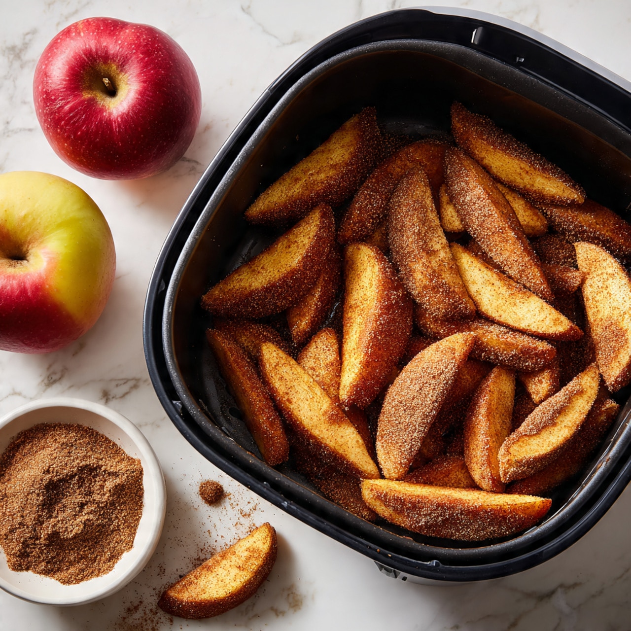 The image shows a black air fryer basket filled with a single layer of apple slices coated in a brown cinnamon sugar mix. The apple slices are arranged close to each other covering most of the basket's surface. Nearby, on a white marbled texture surface, sits a whole red and yellow apple, a small white bowl filled with cinnamon powder, and scattered crumbs and pieces of apple slices. The colors include warm brown tones from the sugar coating, the red and yellow of the apple, and the black basket, all set against the white marbled background. Photo taken with an iphone --ar 4:5 --v 7