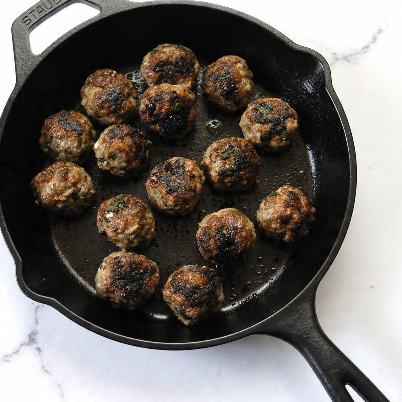 A black cast iron pan holds about twenty small round meatballs, each one browned with crispy dark golden patches on top, showing a textured mix of herbs and greens inside. The meatballs are arranged in a loose pile across the pan’s shiny oily surface. The pan sits on a white marbled surface that is clean and simple. The handle of the pan is visible, labeled with