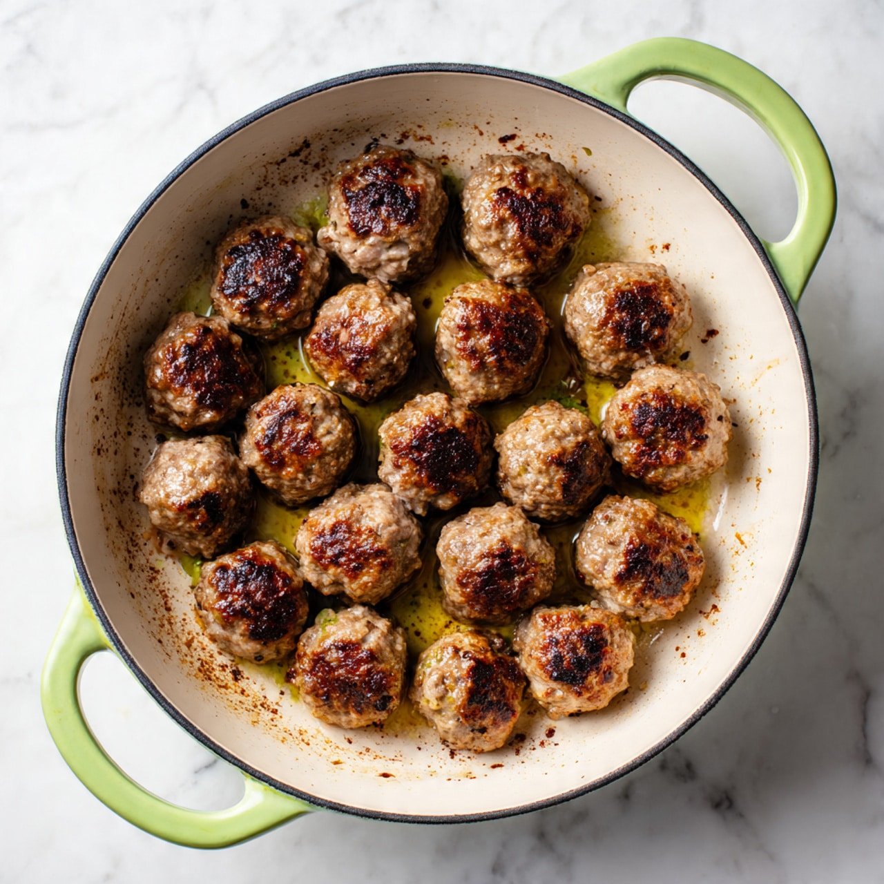 The image shows a white enamel pan with light green handles sitting on a white marbled surface. Inside the pan, there are about sixteen small, browned meatballs spread evenly, each having a dark, crispy outer layer with some visible grill marks and bits of cooked spices around them. The pan’s surface has brown cooked residue and oil spots, giving a rustic look. The meatballs’ rough texture and uneven color shades suggest they are well-cooked on all sides. photo taken with an iphone --ar 4:5 --v 7