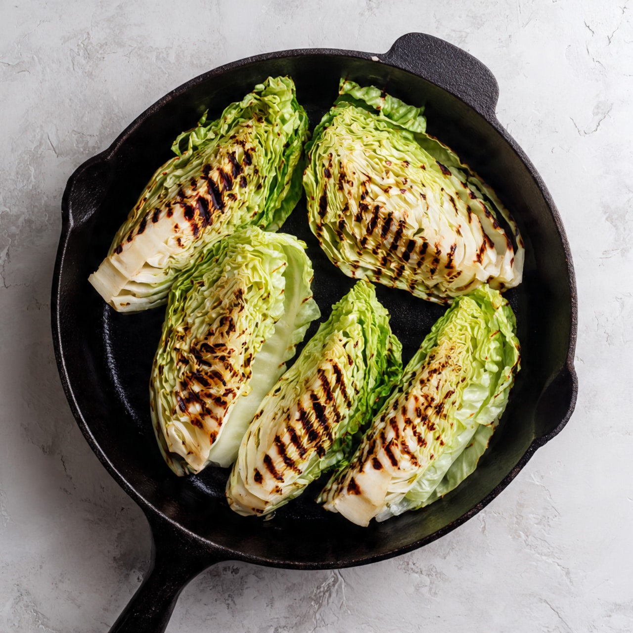 Four grilled wedges of cabbage are arranged inside a black cast iron skillet. Each wedge shows layers of green and white leaves with charred brown grill marks on the cut sides. The wedges are placed spread apart, showing the grill marks clearly against the smooth green outer leaves. The skillet handle is visible at the bottom of the image, and the skillet rests on a white marbled textured surface. The lighting highlights the texture and color contrasts of the cabbage and skillet photo taken with an iphone --ar 4:5 --v 7