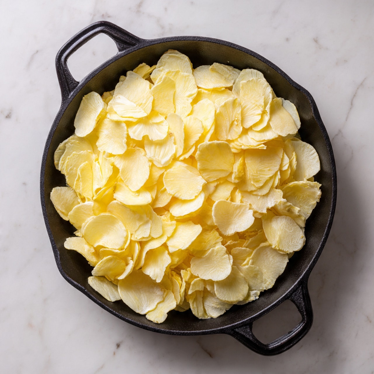 A black cast iron pan filled with one layer of thinly sliced yellow potatoes evenly spread to cover the bottom. The potatoes have smooth edges with some slight texture and natural variations in their pale yellow color. The pan handle points downward, resting on a white marbled surface. Photo taken with an iphone --ar 4:5 --v 7