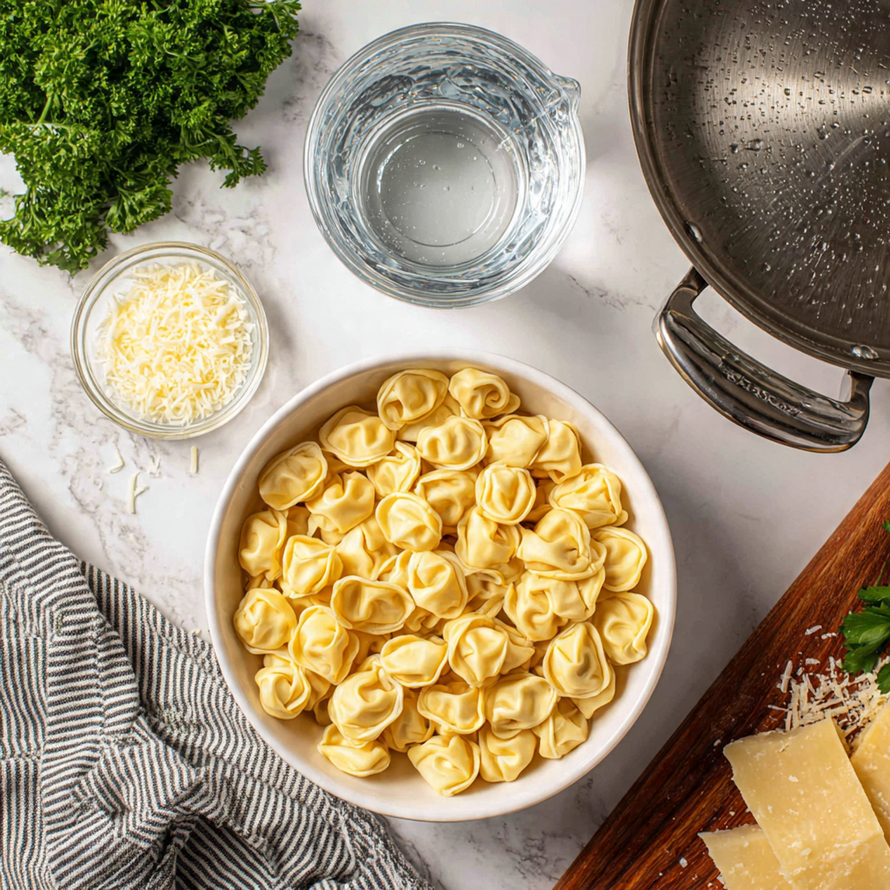 The image shows a white bowl filled with uncooked tortellini pasta at the bottom left, each piece light yellow with a soft, folded texture. Above the bowl is a clear glass measuring cup with water inside, centered on a white marbled surface. To the right of the cup is a large silver metal pan with two handles and a smooth, slightly reflective surface. At the top right corner, there is a wooden board with finely grated and block pieces of pale yellow cheese, some scattered shavings creating a textured area. On the top left, fresh green parsley with curly leaves adds a touch of color, next to a small clear dish of finely minced garlic. A white towel with black stripes lies partially under the bowl on the bottom left. photo taken with an iphone --ar 4:5 --v 7
