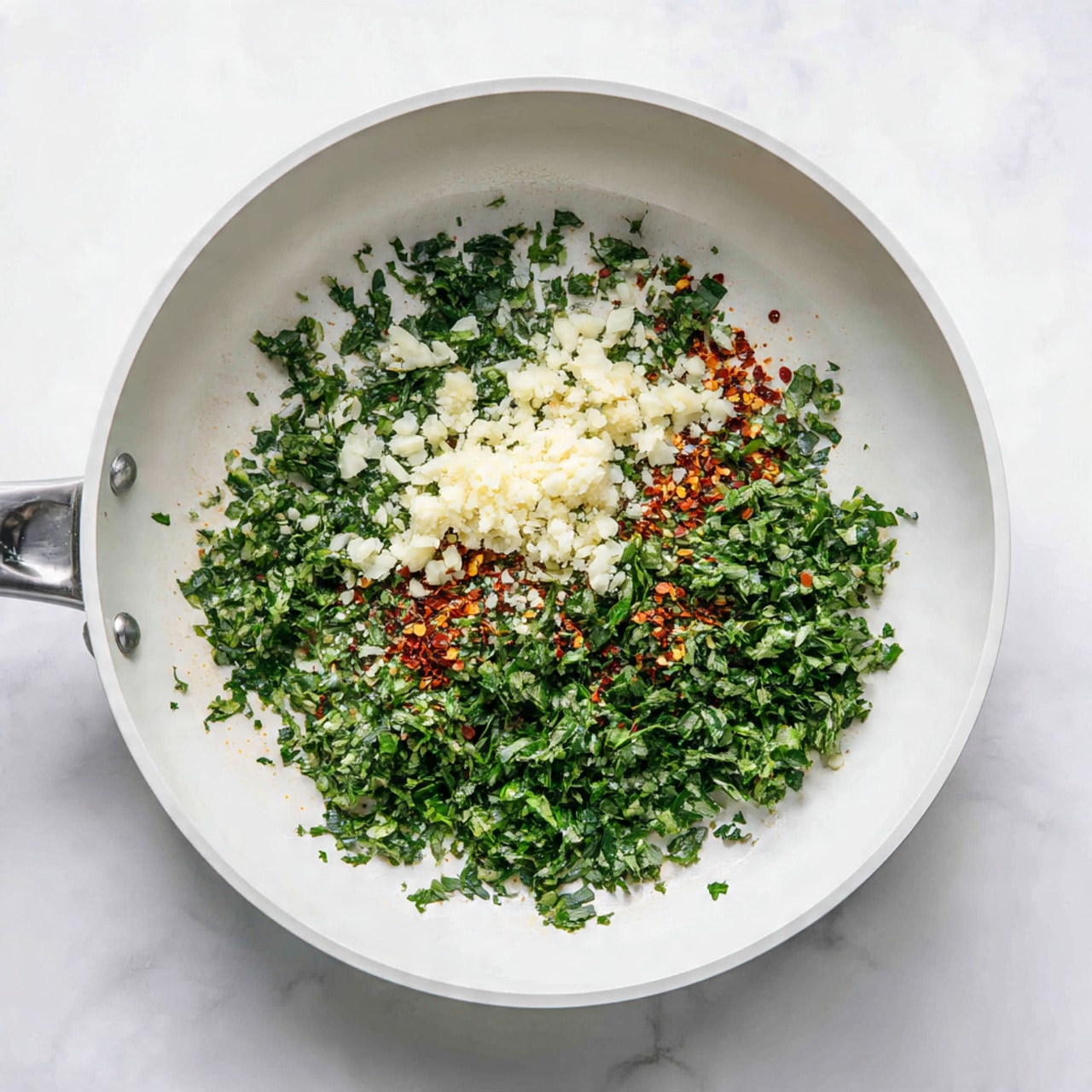 A white frying pan sits on a white marbled surface, filled with three main layers of ingredients. The bottom layer is a spread of bright green finely chopped herbs that cover most of the pan's base. Scattered evenly on top are clusters of off-white minced garlic, adding a rough texture. Around the edges and sprinkled throughout are small patches of red chili flakes, adding a touch of vibrant red color and contrast to the green herbs and pale garlic. Photo taken with an iphone --ar 4:5 --v 7