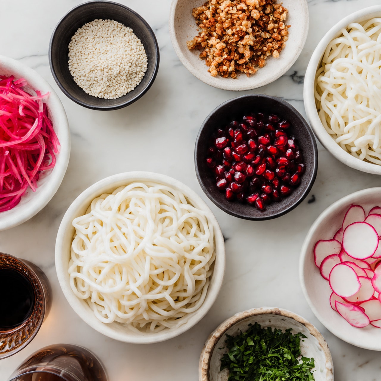 Two white plates on a white marbled surface hold beige pasta arranged in small, loose nests. Around them are five small bowls: one black bowl with white sesame seeds, one black bowl with light brown crunchy topping, one white bowl with thinly sliced pink radishes, one white bowl filled with bright red pomegranate seeds, and one small bowl of chopped green herbs. A glass with a dark sauce is partially visible at the bottom left. photo taken with an iphone --ar 4:5 --v 7