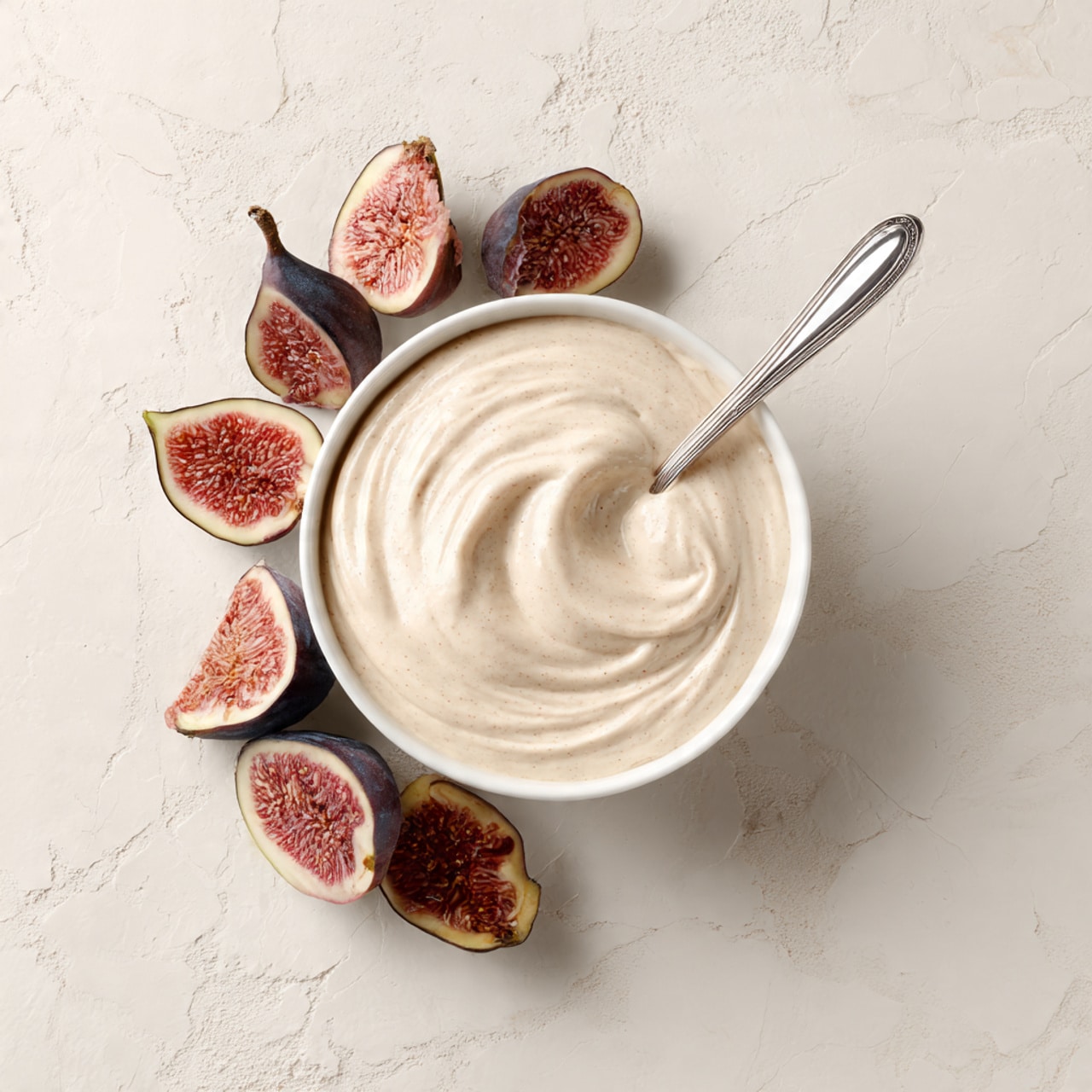 A white bowl filled with a smooth, creamy light beige dip sits in the center of a white marbled surface. A silver spoon is placed inside the bowl, partially submerged in the dip, with the handle resting upright. Around the bowl, there are several halves of fresh figs, showing their dark purple skin and soft, pinkish-red, seed-filled inside, scattered in a loose circle. The whole scene has a clean, fresh look with the contrast of the figs' rich color against the creamy dip and white marble background. photo taken with an iphone --ar 4:5 --v 7