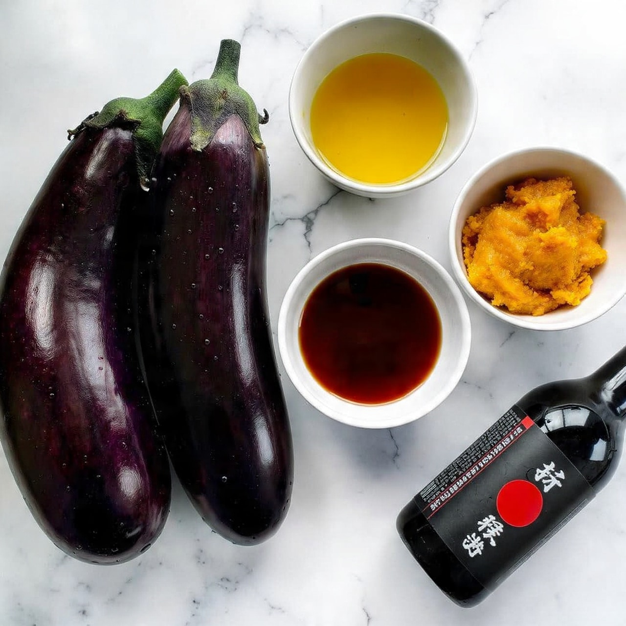 Two large shiny dark purple eggplants with water droplets rest on a white marbled surface. Next to them are three small white bowls: one filled with golden yellow oil, another holding a dark brown liquid, and the third containing thick orange paste. A black bottle labeled