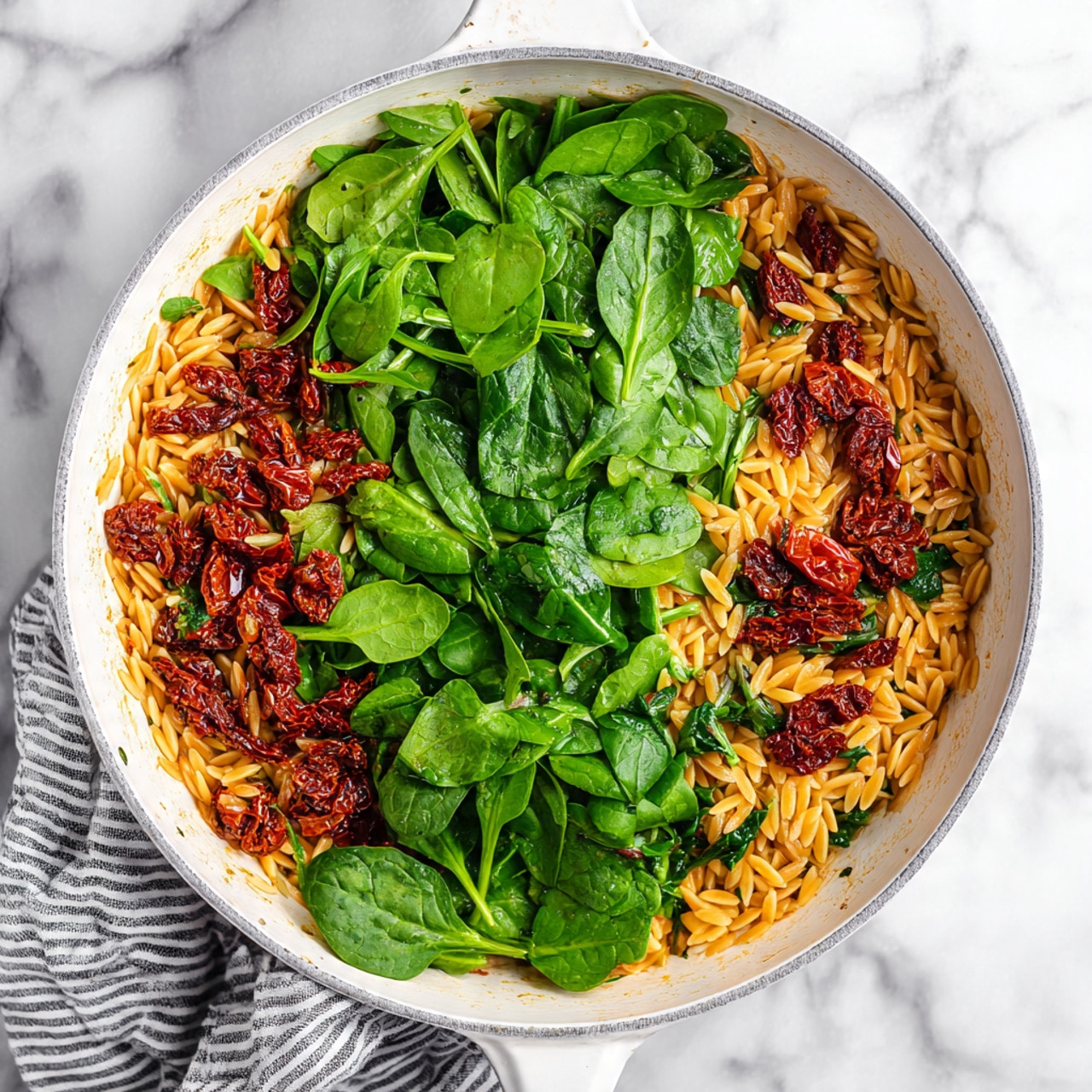 A white pan with a dark rim and handles is placed on a white marbled surface with a striped cloth beside it. Inside the pan, on the left side, there is one layer of cooked orzo pasta mixed with small pieces of sun-dried tomatoes, showing a golden yellow color with darker brown spots. On the right side, there is a layer of fresh, bright green spinach leaves, fresh and whole, covering almost half the pan. The contrast between the warm orzo and the cool green spinach is clear. Photo taken with an iphone --ar 4:5 --v 7
