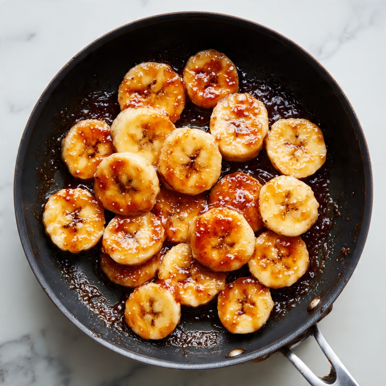 A close-up view of a black frying pan with a flat cooking surface, inside which there are several thick round slices of banana spread out evenly. The pan surface has a layer of dark brown syrup or caramel underneath and around the banana slices, giving it a glossy texture. The pan is set against a white marbled surface background, with no other objects visible. Photo taken with an iphone --ar 4:5 --v 7
