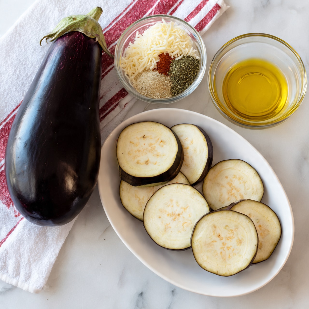 A tall whole eggplant with a dark purple shiny skin lies next to a white cloth with red stripes on a white marbled surface. To the right, there is a small white oval plate holding five thick, round slices of pale cream eggplant with small brown seeds and dark purple edges, spread slightly overlapping. Above this plate is a clear glass bowl with melted butter showing a shiny, yellow texture. Left of the eggplant slices, a large white bowl holds a dry mix with distinct layers: shredded pale beige cheese, light beige breadcrumbs, dried green herbs, red spice powder, and pale tan powder, each sectioned but touching. The photo taken with an iphone --ar 4:5 --v 7