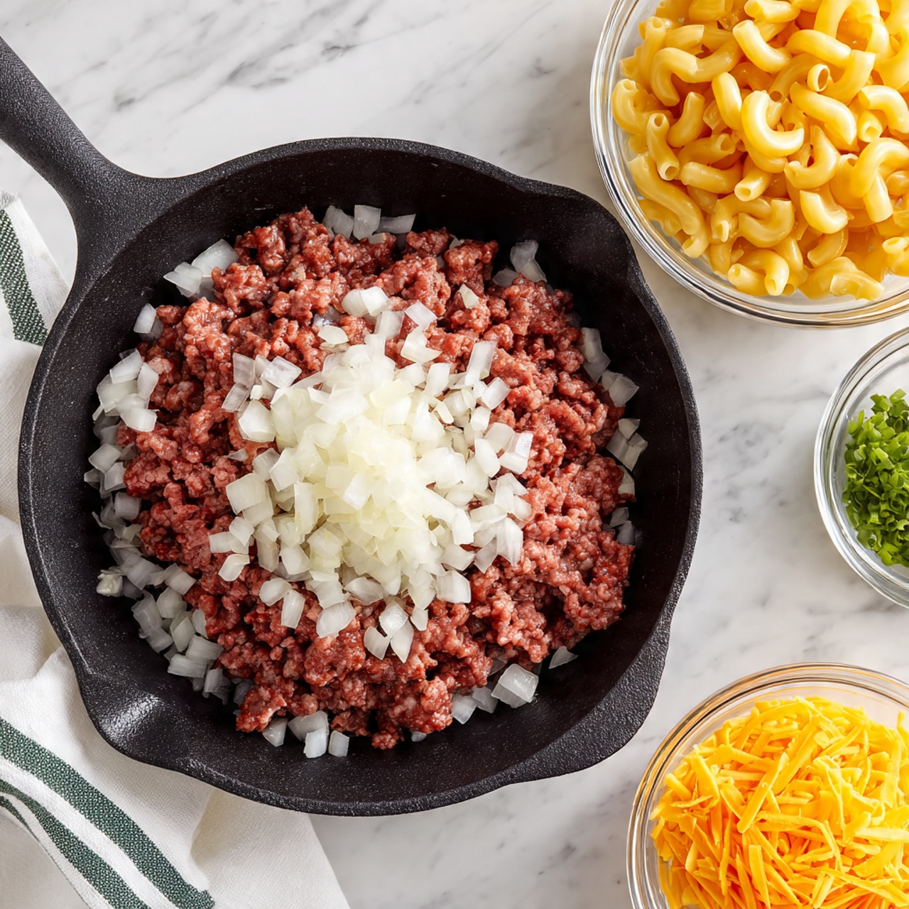 A black cast iron pan sits on a white marbled surface, holding a thick rectangular layer of raw ground beef topped with a layer of white chopped onions scattered unevenly on and around it. Behind the pan, two clear glass bowls rest side by side on the white marbled surface; the left bowl is filled with uncooked yellow elbow macaroni, and the right bowl contains shredded bright orange cheddar cheese. A white kitchen towel with green stripes is placed to the left side of the pan. Photo taken with an iphone --ar 4:5 --v 7