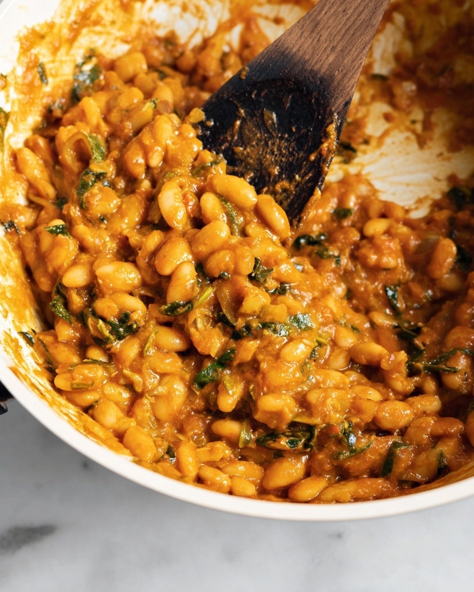 The image shows a close-up of a cooked dish in a white pan with orange sauce coating white beans and small green vegetables mixed throughout. A wooden spoon with a burnt edge is resting in the pan, partially covered with the thick, chunky mixture. The beans and vegetables are mixed evenly, creating a textured surface with glossy, soft orange sauce around them. The background features a white marbled surface. photo taken with an iphone --ar 4:5 --v 7