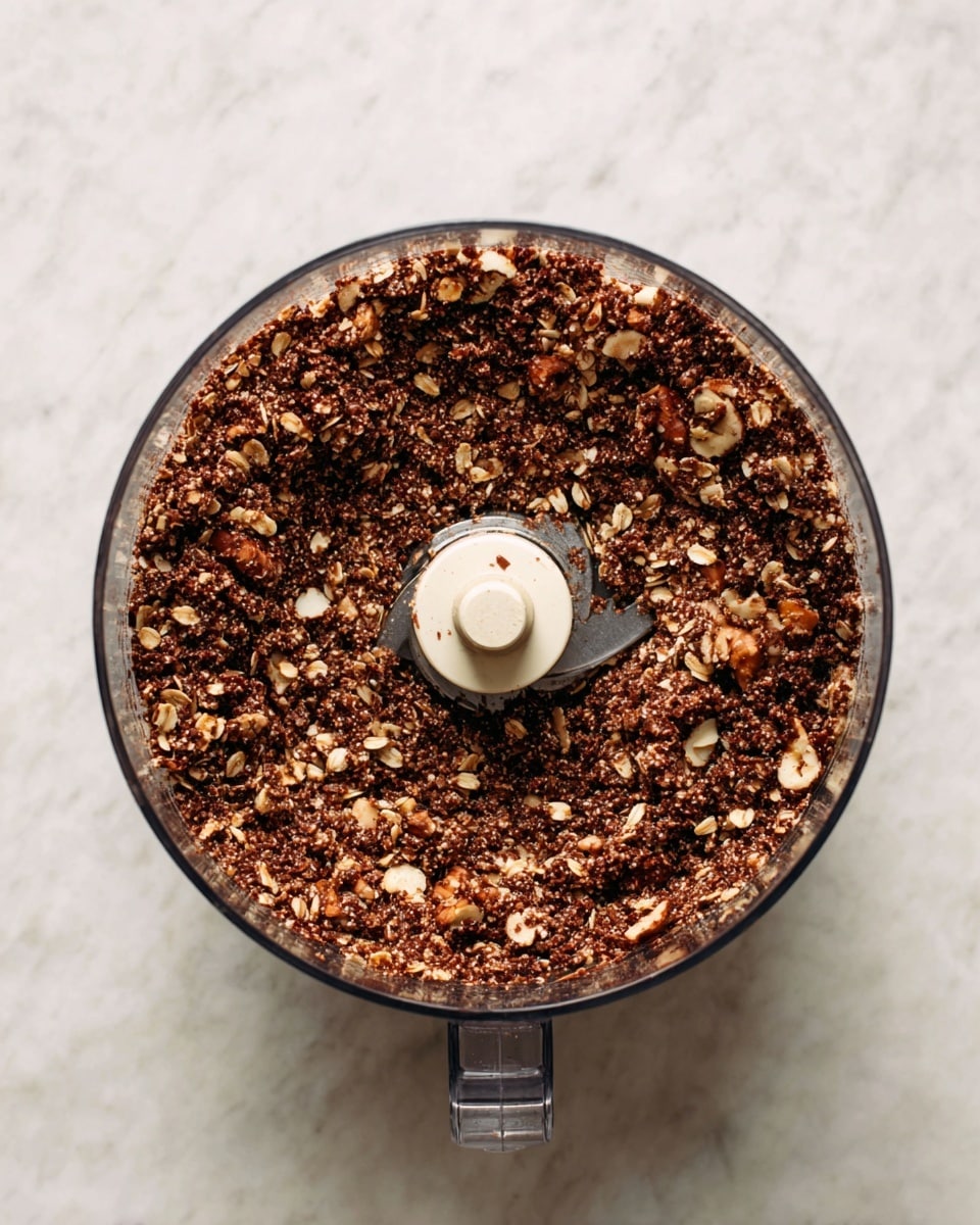 A clear food processor bowl filled with a coarse mixture of oats, nuts, and chocolate, showing a rough and chunky texture with visible small pieces mixed evenly throughout. The mixture is dark brown, with some lighter bits of oat and nut pieces scattered inside. The food processor's blade center is partly visible, surrounded by the mixture, and the whole container sits on a white marbled surface. photo taken with an iphone --ar 4:5 --v 7