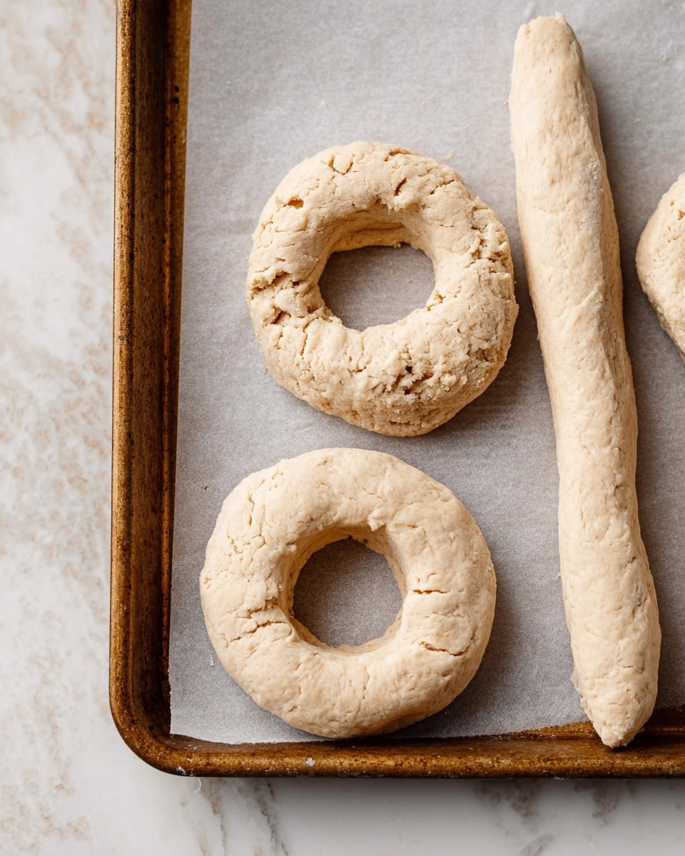 The image shows an old brown metal baking tray lined with light gray parchment paper, placed on a white marbled surface. On the tray, there are three raw dough rings with rough, uneven textures and pale beige color, each shaped into a round ring with a hole in the middle. Next to the rings is a long, thin dough piece, similarly textured and colored, lying horizontally across the tray. The dough looks soft and unbaked, with some cracks and irregularities visible on the surface. photo taken with an iphone --ar 4:5 --v 7