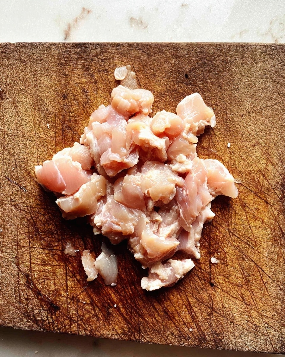 A close-up view of small pieces of raw chicken meat placed on a worn brown wooden cutting board with visible knife marks and scratches. The chicken pieces are pale pink with some white fatty parts, loosely grouped in the center of the photo. The scene is set on a white marbled textured surface. Photo taken with an iphone --ar 4:5 --v 7