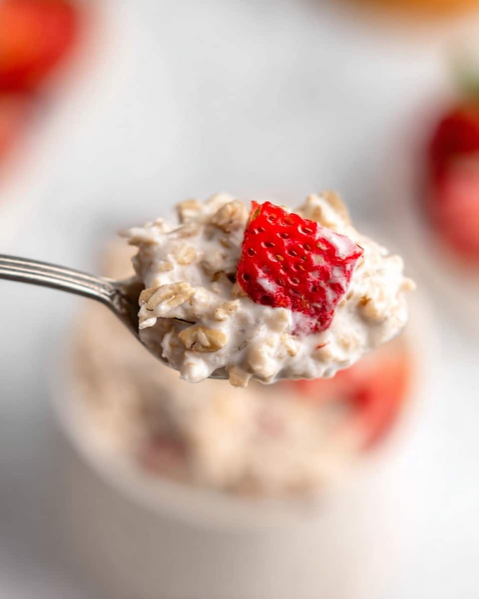A close-up of a silver spoon holding a scoop of creamy oatmeal mixed with visible oats, and a bright red chunk of strawberry on the side, set against a blurred white background with hints of red and orange colors. The texture of the oatmeal looks soft and slightly thick, with small pieces of oats and seeds visible throughout. In the out-of-focus background, there's a white container with red strawberry pieces inside, placed on a white marbled surface. photo taken with an iphone --ar 4:5 --v 7