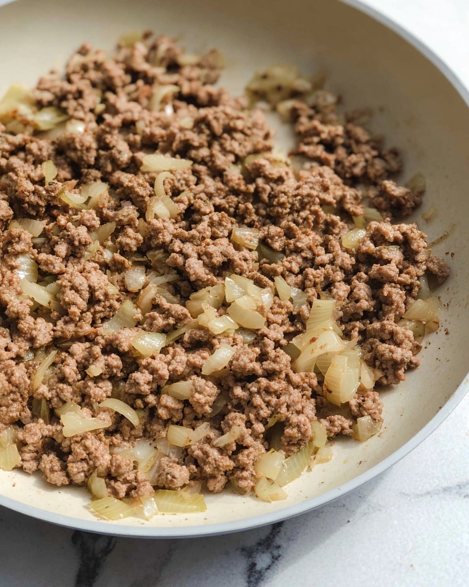 A close-up view of cooked ground meat mixed with small, translucent pieces of cooked onions inside a white frying pan, showing rough texture and light brown and pale yellow colors. The pan rests on a white marbled surface, with soft natural light highlighting the food's details. photo taken with an iphone --ar 4:5 --v 7