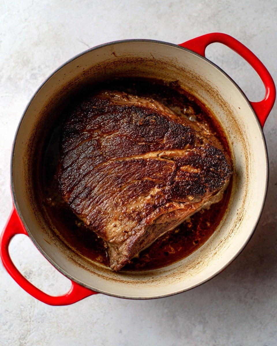 A large piece of browned meat sits in the middle of a white pot with bright red handles, the meat showing a rich dark brown sear with textured surface and some glistening juices around it. The inner walls of the pot show signs of cooking with a slightly browned ring near the top, while the white marbled surface is visible underneath the pot. The image is clear and bright, taken from above, focusing on the cooked meat inside the pot photo taken with an iphone --ar 4:5 --v 7