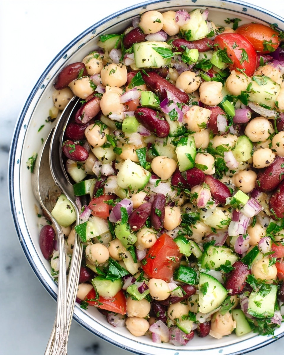 A white bowl with a thin dark blue rim holds a colorful bean salad mixed with chopped cucumber, tomato, red onion, and fresh green parsley. The salad combines three types of beans: creamy white, light beige chickpeas, and dark red kidney beans, all mixed evenly throughout. Small bits of chopped green celery add more texture. The ingredients appear fresh and are sprinkled with dried herbs, giving a speckled green and black effect across the vegetables and beans. A pair of vintage-style metal salad spoons rest inside the bowl on the left side. The bowl sits on a white marbled surface photo taken with an iphone --ar 4:5 --v 7