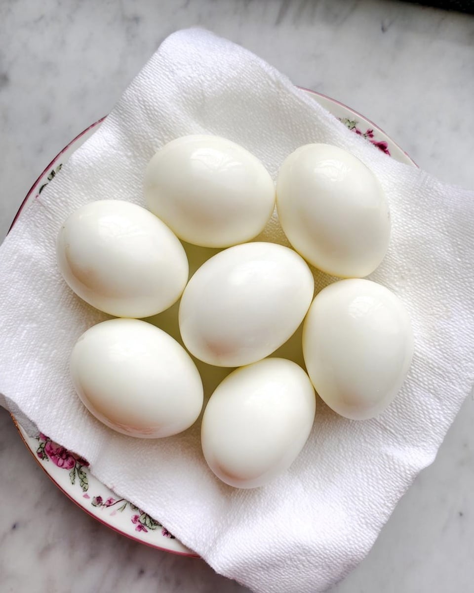 Seven peeled boiled eggs are placed on a white plate with a light floral pattern, which is covered with a white paper towel. The eggs are smooth and shiny, with a white color and oval shape. The plate sits on a white marbled surface. Photo taken with an iphone --ar 4:5 --v 7