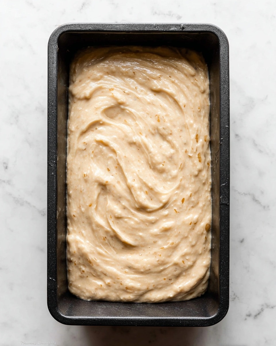 The image shows a single rectangular black baking pan filled with a thick, creamy batter that has a light beige color with small specks and chunks visible throughout. The surface of the batter is smoothed out with gentle swirls and waves, creating a soft texture on top. The pan sits on a white marbled surface, highlighting the contrast between the dark pan and the pale batter inside. photo taken with an iphone --ar 4:5 --v 7